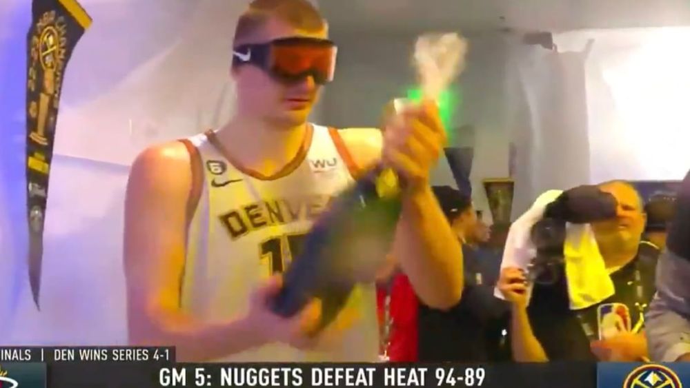 Nikola Jokic in the Nuggets locker room in a Nuggets jersey, with goggles on, half-heartedly shaking a bottle of champagne. He is neither smiling nor frowning, he is completely emotionless