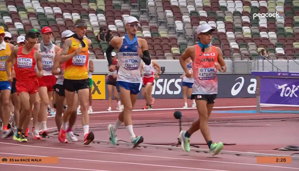dozens of competitive racewalkers are doing their thing. they're in a track-and-field stadium but nobody is in the stands. they simultaneously look like they're exerting a lot of energy and yet chilling at the same time. the guy in front has both of his feet off the ground which means he is CHEATING 