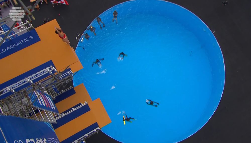 a woman stands on a platform, facing backwards, her ankles over the edge. many many many feet beneath the platform is a circular pool filled with people in fins and scuba gear