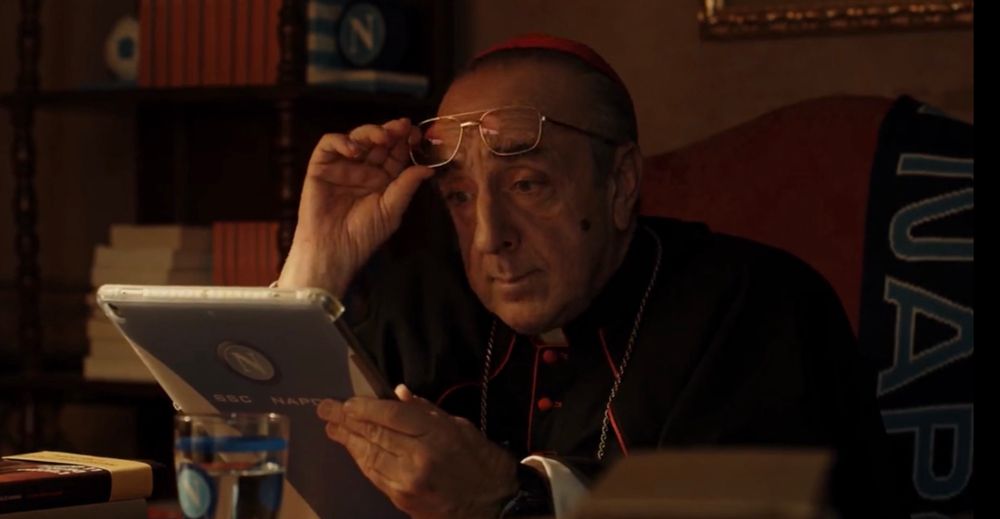 A Cardinal reading an iPad with a Napoli case, on a chair with a Napoli scarf, in front of a table with a Napoli water glass, and a shelf with Napoli books 