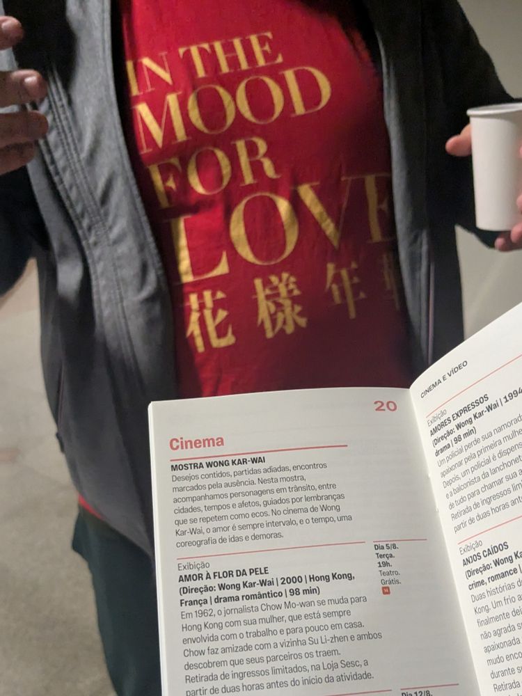 tronco de um homem gesticulando com um copo na mão. ele usa uma camisa vermelha com estampa do filme "in the mood for love" (tradução: amlr à flor da pele) e uma jaqueta cinza. em frente a ele, eu seguro um guia de programação aberto na página do cinema, onde tá tendo mostra wong kar wai e, hoje, será exibido "in the mood for love"

o conteúdo do guia:
"MOSTRA WONG KAR-WAI

Desejos contidos, partidas adiadas, encontros marcados pela ausência. Nesta mostra, acompanhamos personagens em trânsito, entre cidades, tempos e afetos, guiados por lembranças que se repetem como ecos. No cinema de Wong Kar-Wal, o amor é sempre intervalo, e o tempo, uma coreografia de idas e demoras.

Exibição

AMOR À FLOR DA PELE

(Direção: Wong Kar-Wai | 2000 | Hong Kong, França | drama romántico | 98 min)

Em 1962, o jornalista Chow Mo-wan se muda para Hong Kong com sua mulher, que está sempre envolvida com o trabalho e para pouco em casa. Chow faz amizade com a vizinha Su Li-zhen e ambos descobrem que seus parceiros os traem. Retirada de ingressos limitados, na Loja Sesc, a partir de duas horas antes do início da atividade."