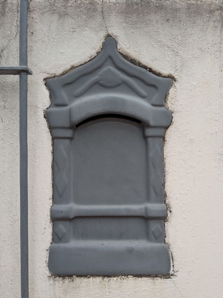 caixa de correio cinza em um muro branco. ela tem formato de palacete como as clássicas, mas sem os detalhes que elas geralmente possuem. a tinta escolhida para sua pintura é fosca, e dá a impressão de que a caixa é feita de argila ou massinha.