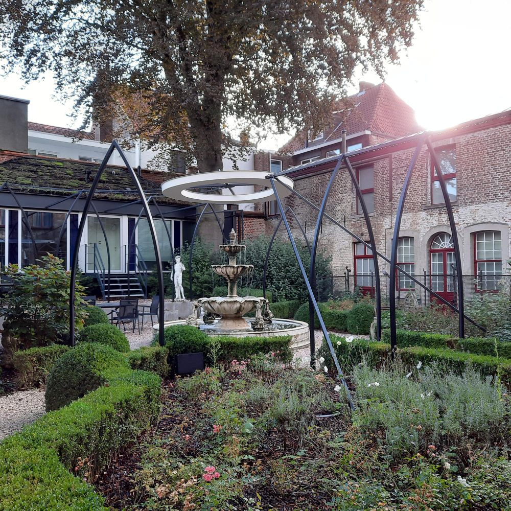 Blumenbeet mit Buxbaumhecke  in einem Hinterhof. Darüber eine runde Lichtinstalltion. Hinter dem Beet ist ein Springbrunnen und eine weiße Statue.
