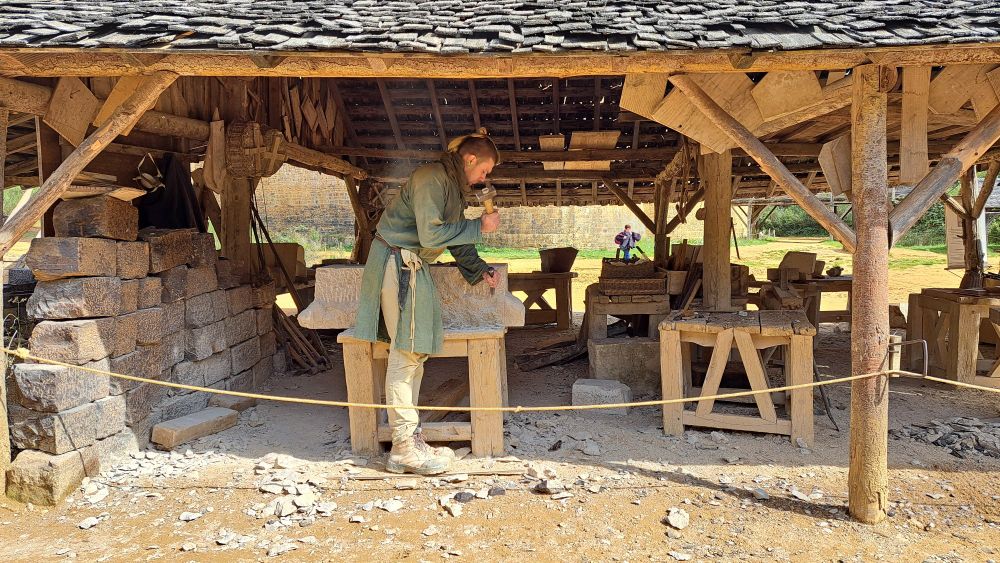Ein Mann in einer grünen Tunika und heller Hose steht in einer offen Hütte und bearbeitet einen großen Steinblock mit Hammer und Meißel