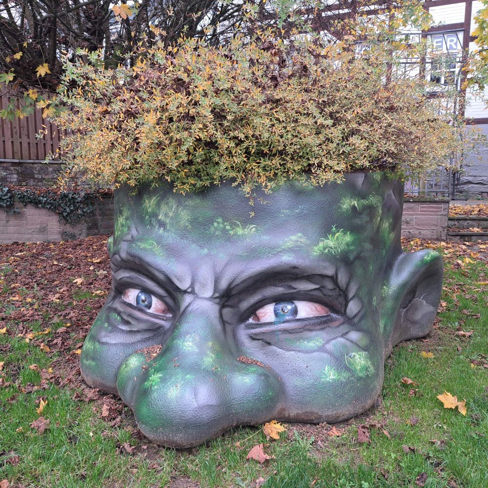 Sehr großer Pflanzentopf in dem des Gesichts eines grünen Trolls oder ähnliches.  Die gelb grünen Sträucher bilden die Haare.
