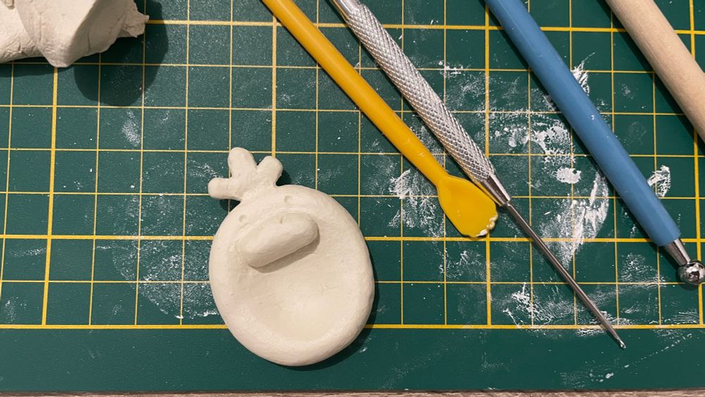 A clay worry stone, unpainted, with psyduck features, next to tools for clay shaping