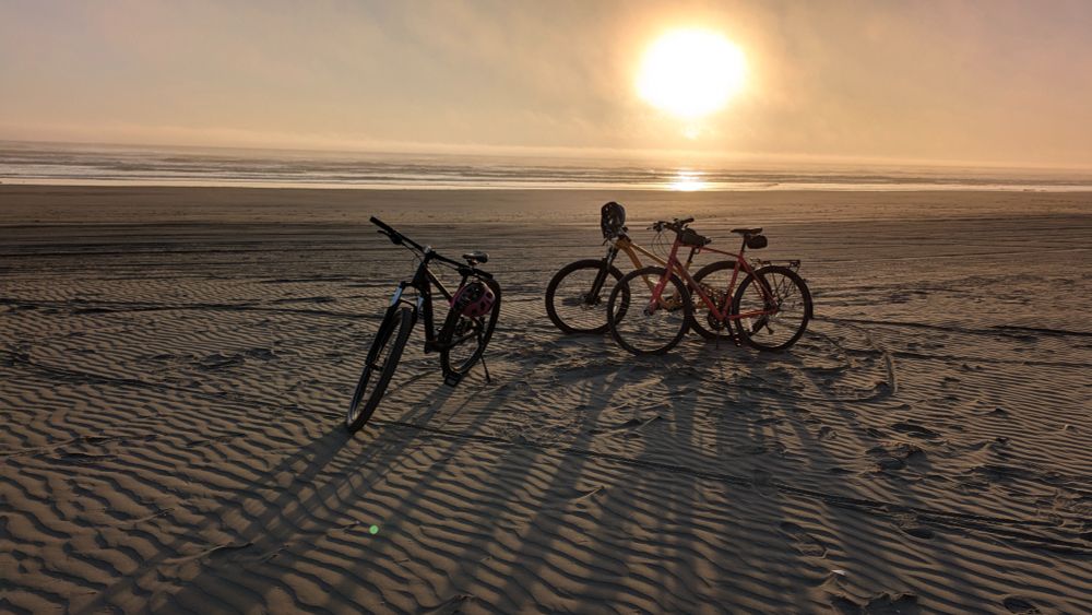 Three bikes on a beach, water and waves in the background with a hazy sun close to setting.
