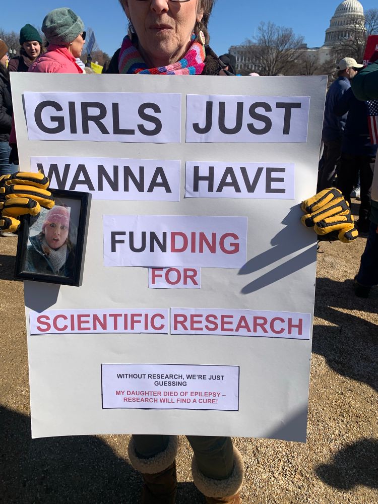 A woman holds a sign that reads, “Girls Just Wanna Have Funding for Scientific Research” while holding a photo of her daughter, who died from epilepsy. 

The sign further reads, “Without research, we’re just guessing. My daughter died of epilepsy. Research will find a cure!” 

