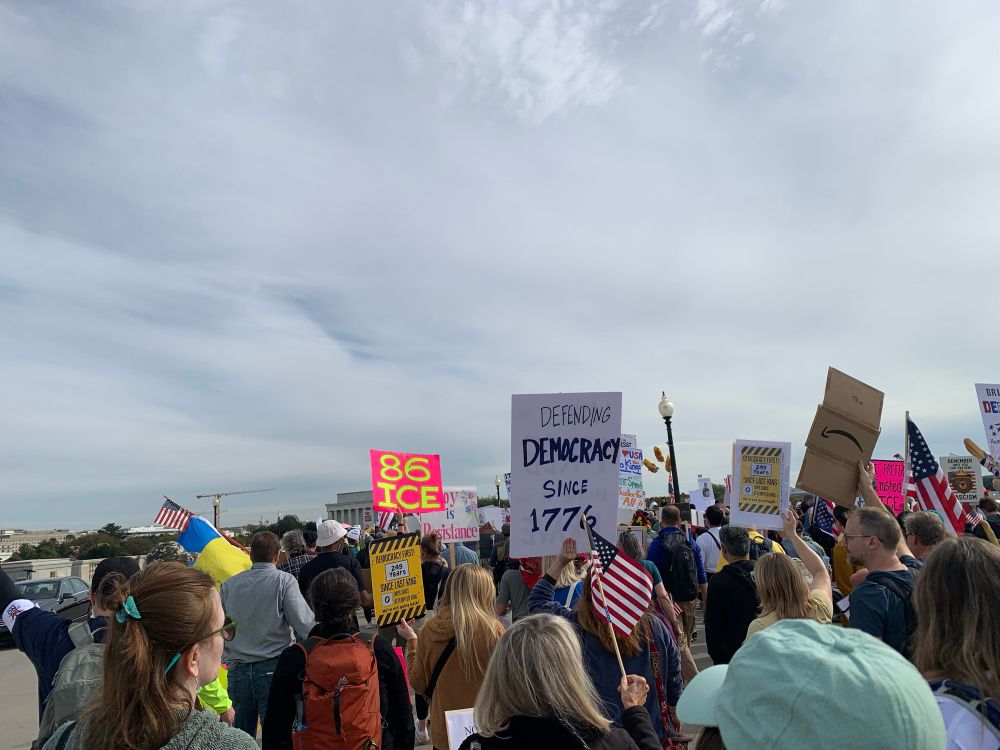 A large crowd of people marching across Memorial Bridge. The Lincoln Memorial is just barely visible ahead of them. Signs include, “86 ICE” and Defending Democracy Since 1776.” 