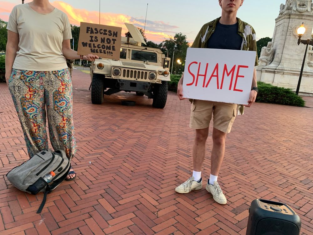 Two protesters standing in front of the National Guard with signs that say, “Shame” and “Fascism Is Not Welcome Here.” 
