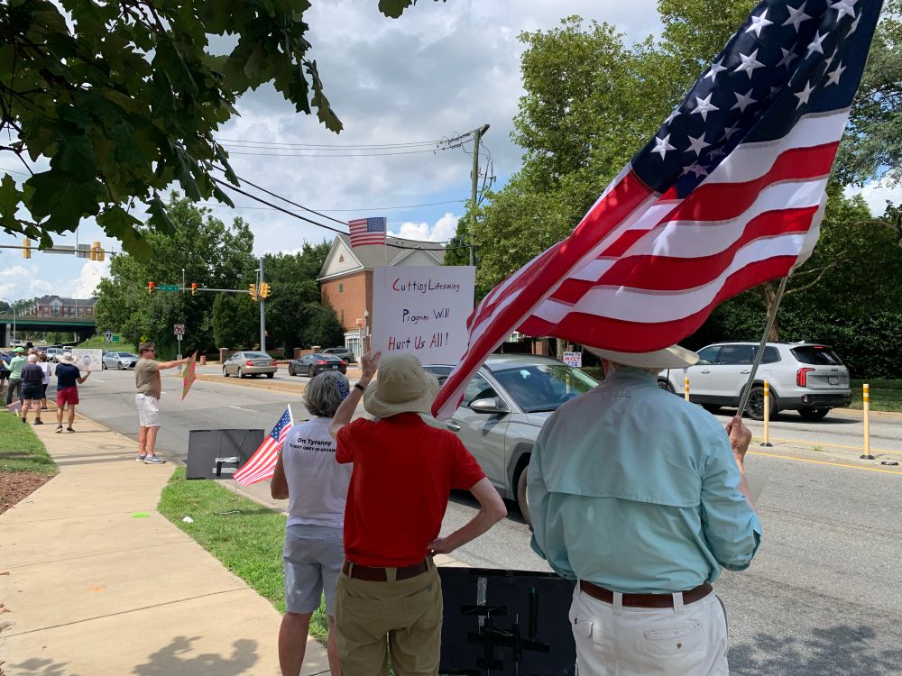 Three #TeslaTakedown protesters. One holds an American flag whipping in the wind. The person next to them holds a sign that says, “Cutting Life-saving Programs Will Hurt Us All.”
And the third wears a t-shirt that says, “On Tyranny, Do Not Obey in Advance” while holding a small American flag. 