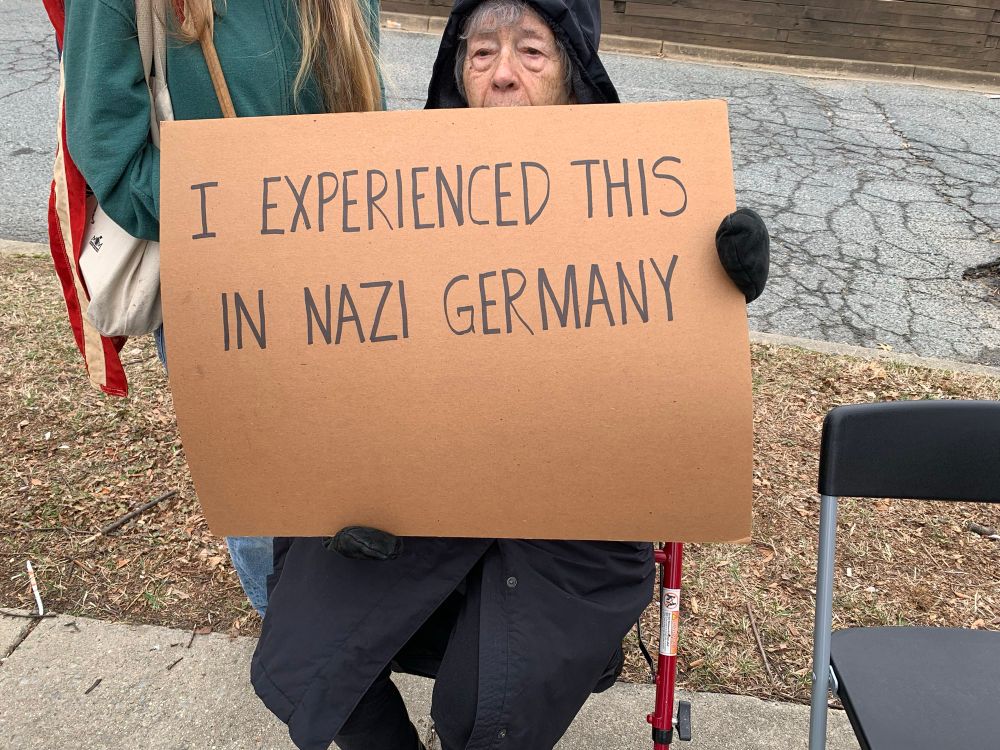 An elderly woman, her face partially obscured, sits on a chair with a sign that says, “I experienced this in Nazi Germany.” 