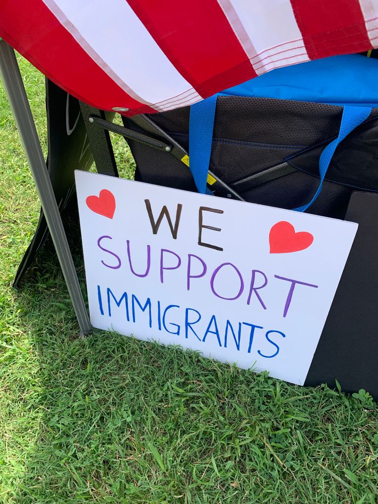 A sign that says, “We Support Immigrants,” with a red heart on either side. The edge of an American flag can be seen at the top of the photo. 