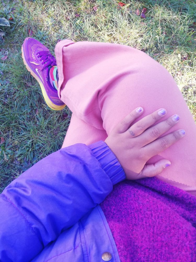 .photo prise de haut - jambes croisées, main gauche se reposant sur la jambe droite. pantalon saumon, pull fuchsia lurex pailleté, doudoune violette, baskets orange et fuschia, chaussettes multi-colores. sur fond de gazon. 