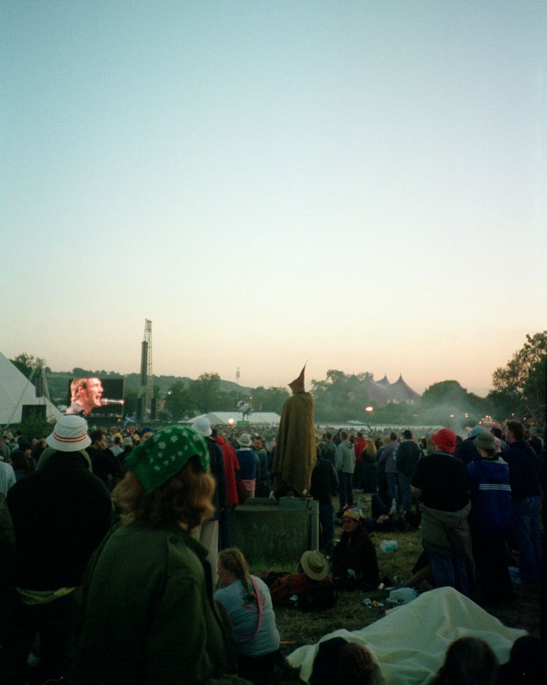 The crowd at Glastonbury 2003 watch David Gray perform as the sun sets. In the centre of the image, a young man in a cloak with a pointed hat stands on top of a drain to get a better view.
