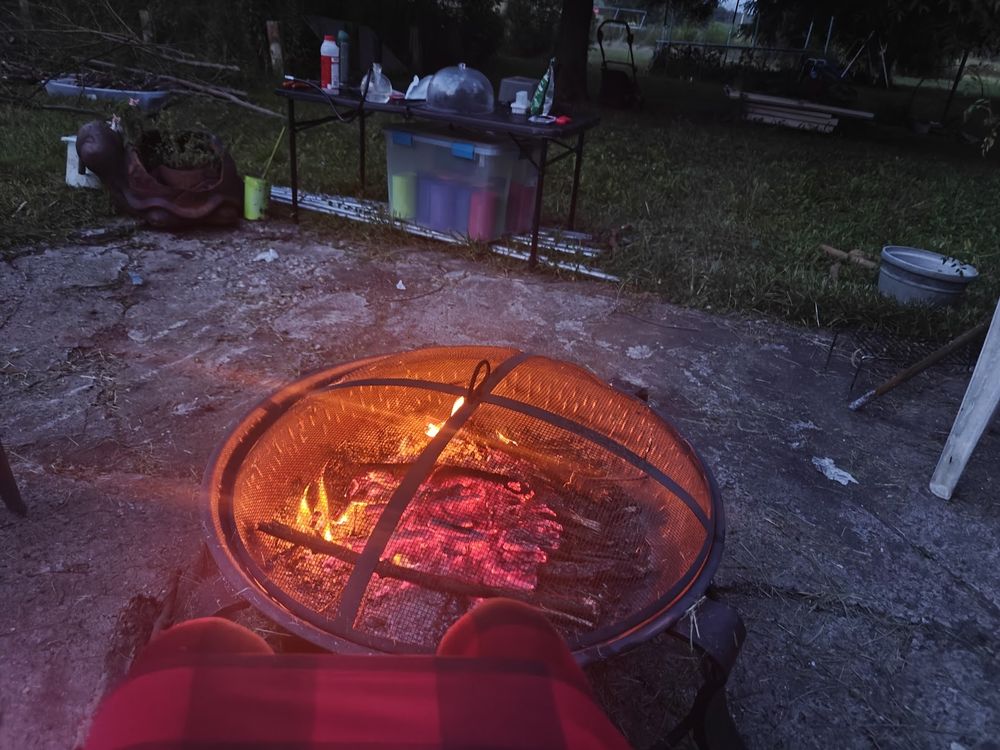 A round, metal mesh-covered fire pit is positioned on a cracked concrete patio, with glowing embers and small flames visible inside. Me wearing red and black plaid pajama pants relaxing. Behind the fire pit, a black metal folding table holds on the table, a bottle of milos, and my orb bong. Under the table is a clear plastic storage bin containing various colors of spray painted tin cans. To the left, our turtle planter with green foliage rests on the ground