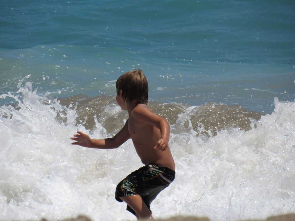 Photograph by Laura Williams of a young boy with sun-kissed skin and wet, tousled hair runs through the foamy waves at the beach. He is shirtless and wearing black swim trunks with a tropical pattern. His arms are outstretched as he looks back toward the crashing surf, mid-motion, with the blue ocean stretching out behind him. The scene captures the energy and excitement of playing in the waves on a sunny day.
