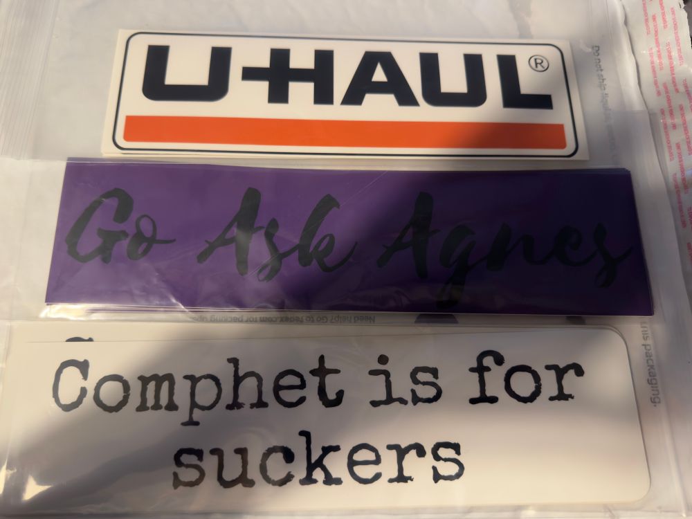 A photo of three bumper stickers in ziplock bags. The top one is the u-haul logo, the second is purple and says “go ask Agnes” in black text, and the third says “comphet is for suckers” in black on a white background.