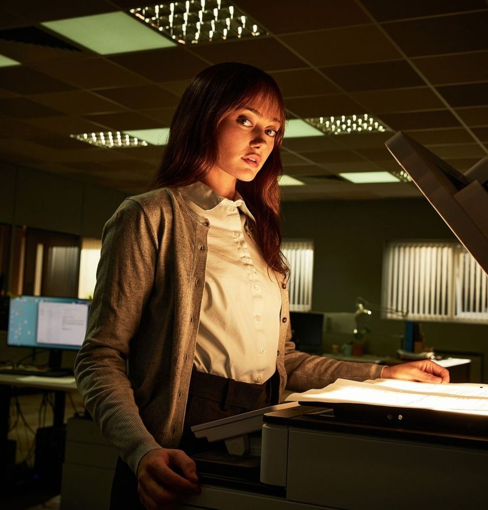 promo image of ella purnell as rhiannon lewis in the television series "sweetpea" (2024). she's using a photocopier in a dark office and looking directly at the camera.