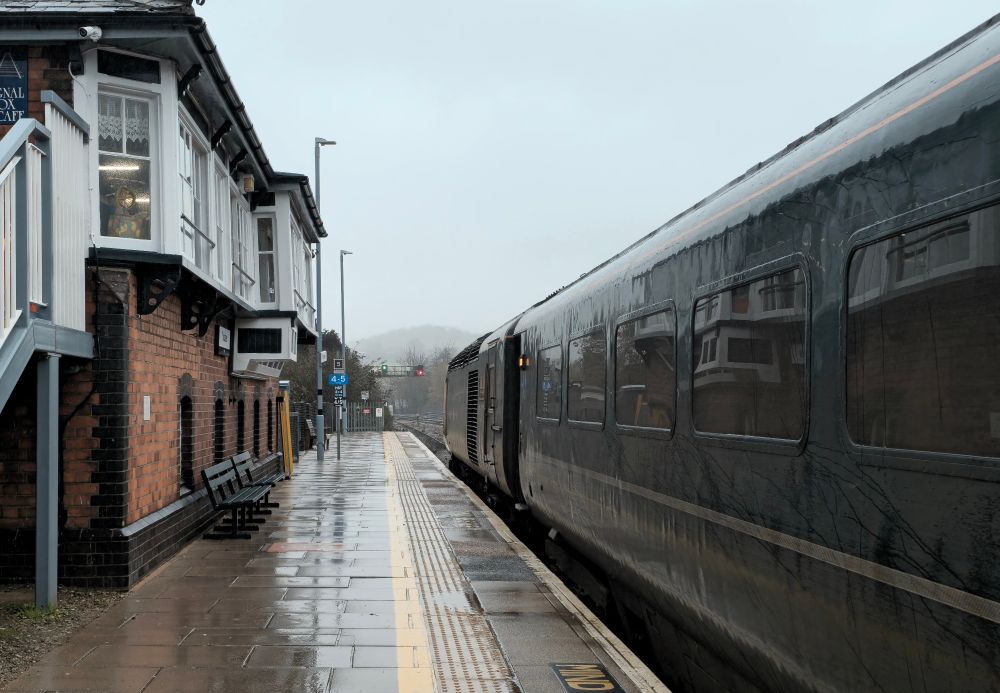 Photo shows a HST next to the signal box at Totnes station. The front power car and carriage is seen in the shot. The signal box is converted into a café. The weather is overcast and wet with poor visibility.