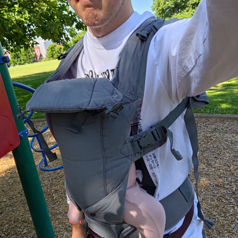 A man wearing a Sniper Zone's Fetish Zone shirt and wearing a baby in a front carrier.