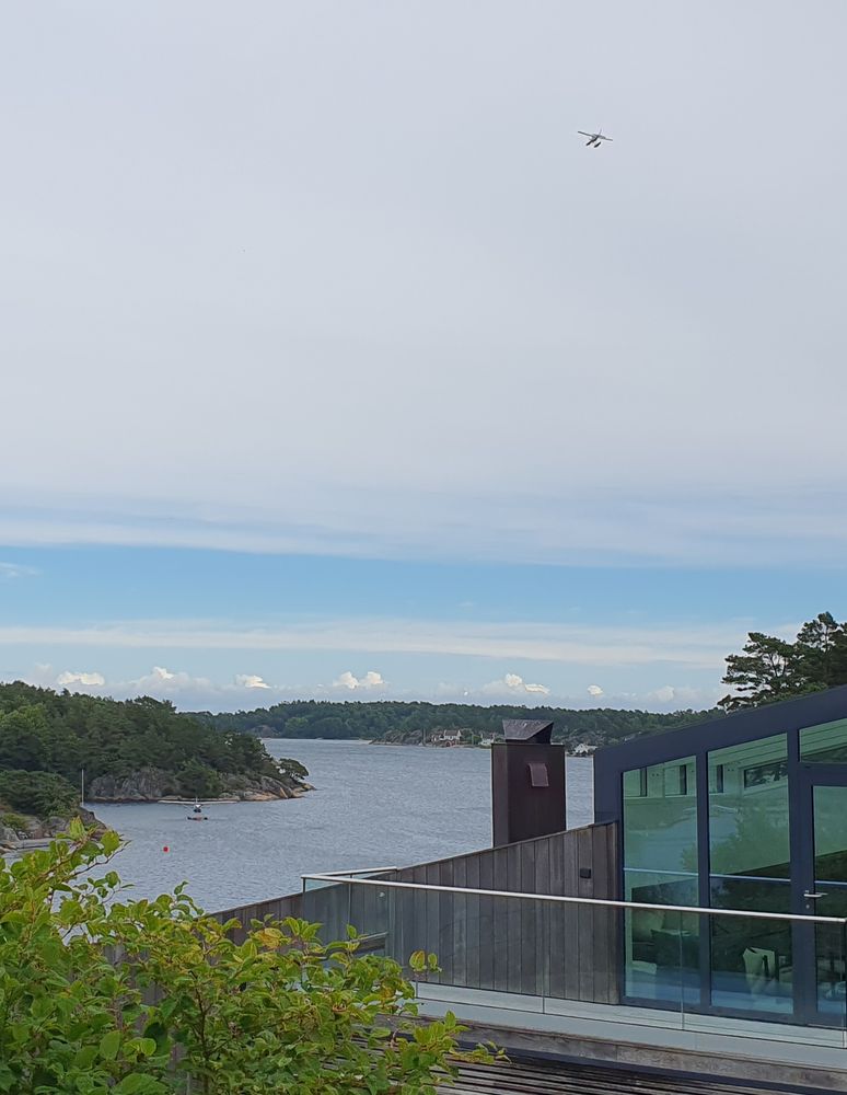 A house in the foreground, a fjord behind it. In the top right a seaplane is heading away, another seaplane is riding at a buoy in the bottom left.
