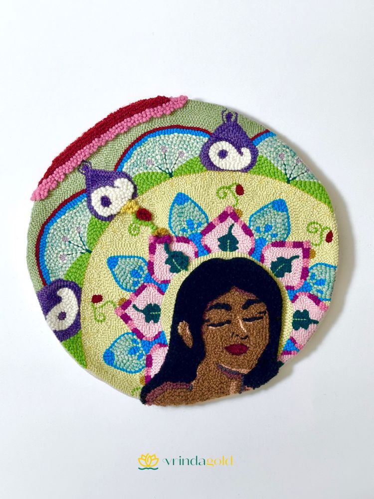 Photo of a punch needle wall art piece. The art piece features a woman with her eyes closed, surrounded by a backdrop of a colourful floral mandala. The word “dream” is incorporated into the mandala. Through the use of reverse and forward punch needling, one instance of the word “dream” is emphasized. 