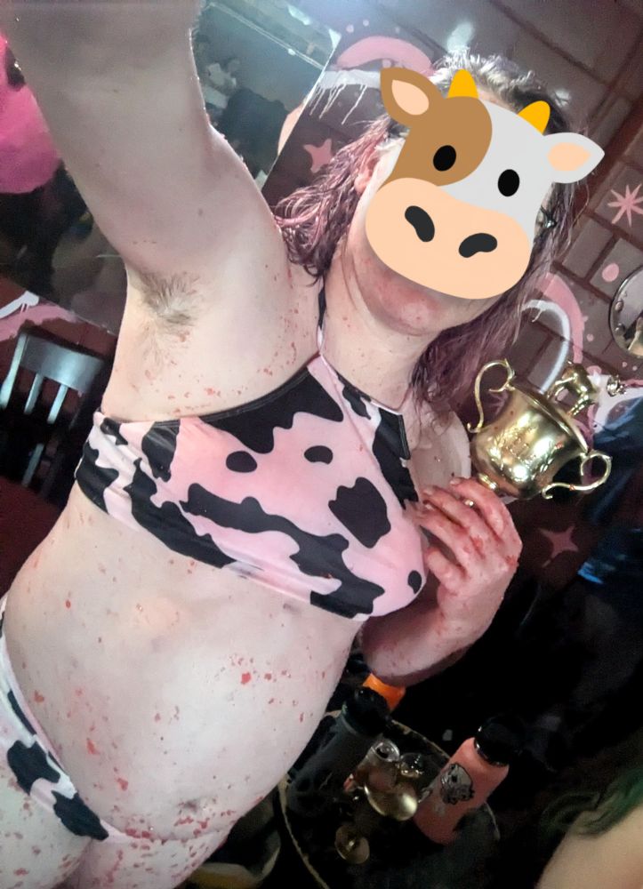 Maggie standing in front of a painted brick wall and mirror. She's wearing a cow print bikini stained pink  and covered with bits of red jello. She's holding a small gold trophy with a goat on top. The cow gave emoji covers her face