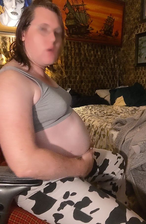 Maggie is sitting in a desk chair, in profile. She's wearing a ribbed grey brallete and cow print jeans. Her belly is sticking out significantly more than usual and she's cupping her under belly in one hand. 