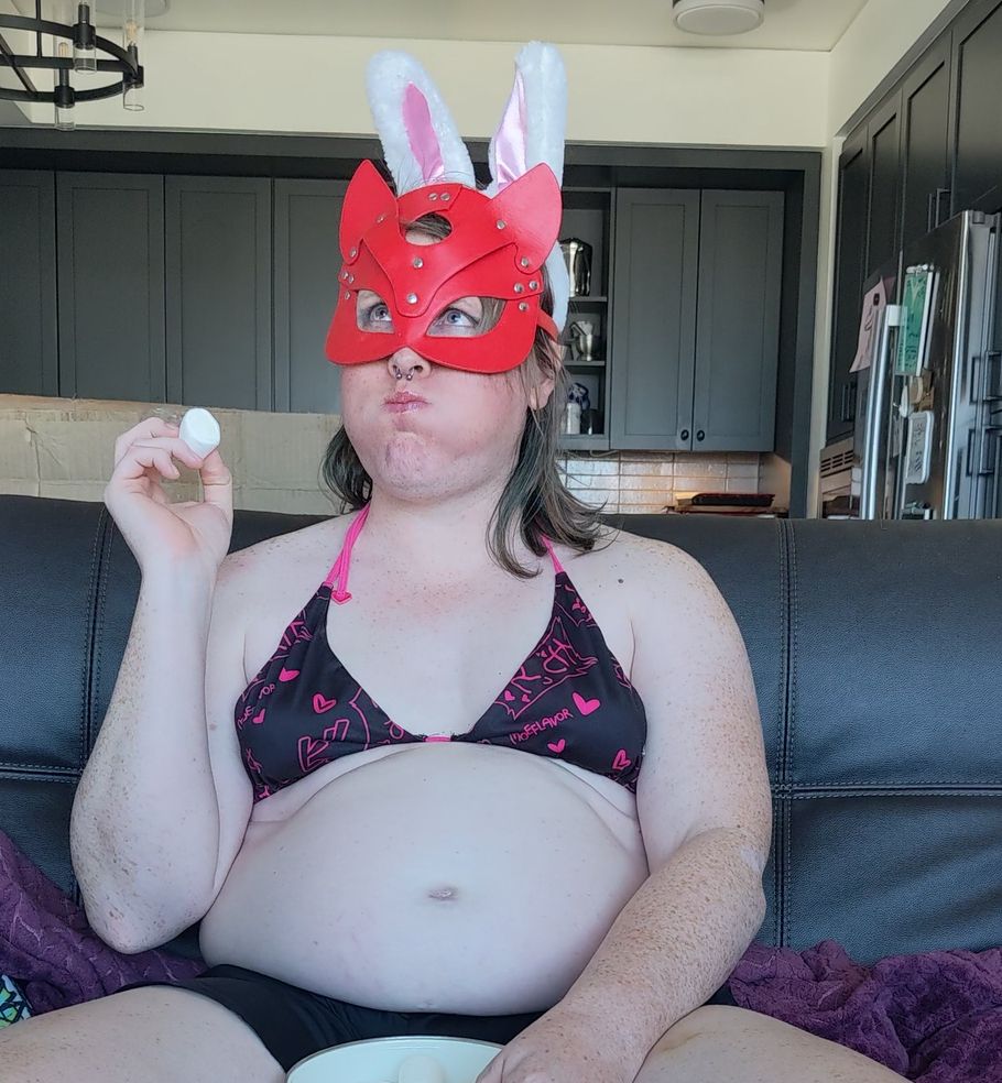Maggie is sitting on a leather couch. She's wearing a black and pink bikini top, a red leather cat mask, and white bunny ears. She has a marshmallow in one hand, and her cheeks are puffed out. Her belly is looking big, with a hint of stretch marks on the lower bit. there's a hint of underboob roles. 