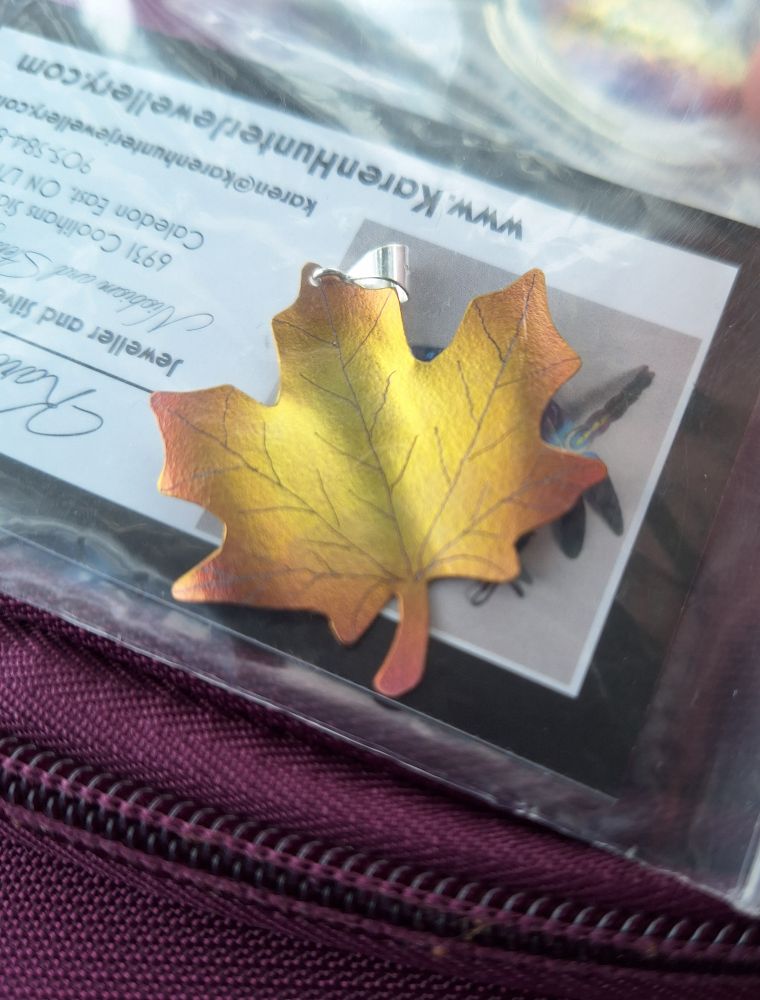 A nobium pendant in the shape of a maple leaf. The leaf is done in shades of gold and copper, with the veins of the leaf traced lightly in black