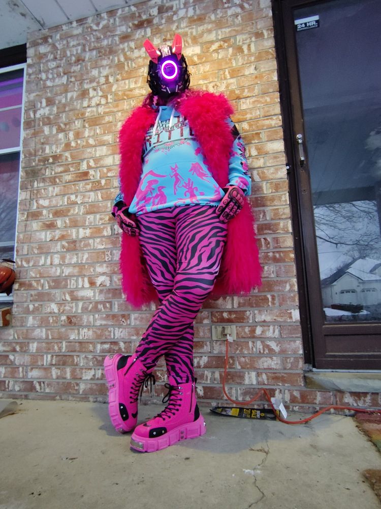 Friend Titan wearing a mask with a pink ring light and pink bunny ear like horns, a pink furry long vest, pink and black tiger striped pants, pink and black boots with chunky soles. They also have a blue crowcorpse hoodie on that says "certified inorganic 100% critter". 

They are leaning casually against a brick wall, thumbs in the pocket of the hoodie