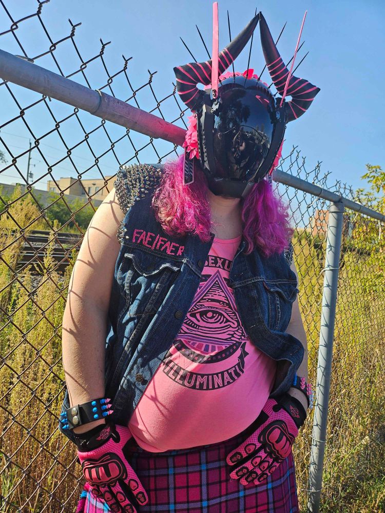 A person wearing a mask with a single featureless dark reflective plate at the front, black horns with pink lines on the sides. The person is also wearing a punk jean best with studs, a pink and blue tartan kilt, bright pink tank top, pink gloves.