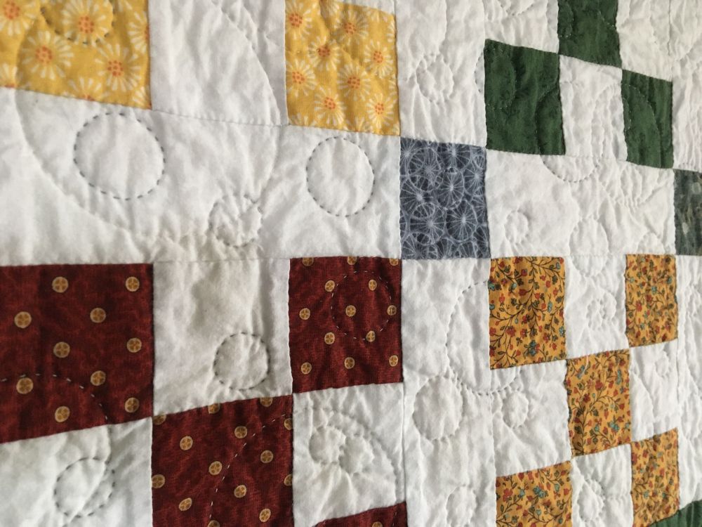 Close-up of a small quilt. It's a kind of checkerboard quilt of white with squares of burgundy, green, blue, and yellow. The quilting itself doesn't follow the squares of fabric, but it is just a mess of circles of various sizes, some overlapping. Kind of whimsical. 