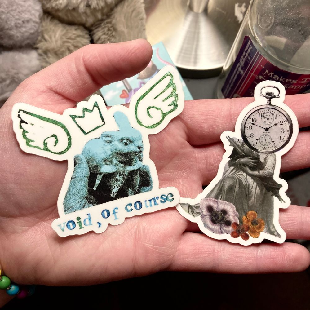 a pair of vinyl stickers held up in the palm of a hand.

sticker on the left: a blue sticker of a hand holding up a ceramic bunny statue. there are block printed wings and a crown around it. across the bottom, block printed letters read "void, of course" in blue ink.

sticker on the right: a stopwatch-headed figure wearing a long white dress cradles a person on a stretcher, with white, red, and yellow flowers at their feet. 