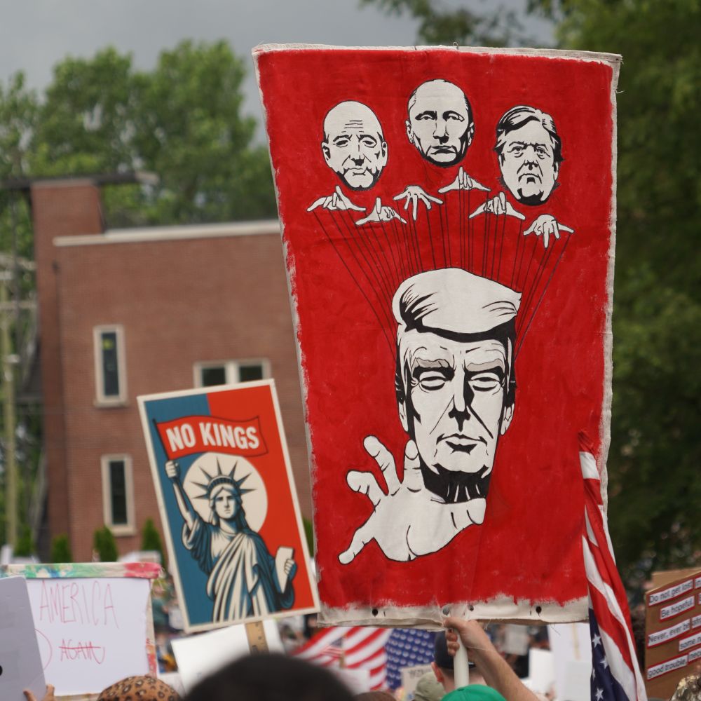 Protest sign with what appears to be puppet masters holding strings over Donald Trump. Next to it is a sign depicting the Statue of Liberty holding a flag that reads "No Kings"