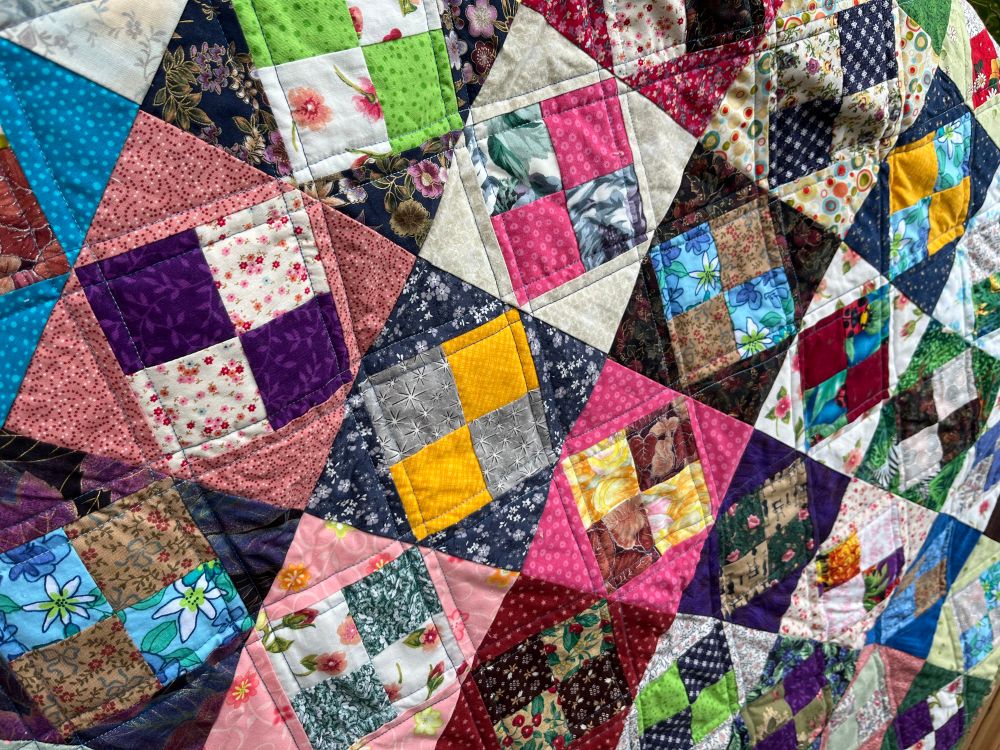 Close up of a scrap quilt made of four patch squares in a larger square. Quilt is machine quilted in straight lines which compliment the lines of the fabric seams. 