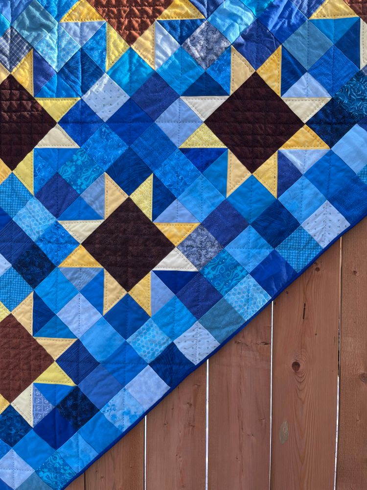 Quilt hanging on a fence made of sunflowers with brown centres and scrappy yellow petals against a scrappy blue background. Quilt is hand quilted with a small grid pattern on the brown centres. 
