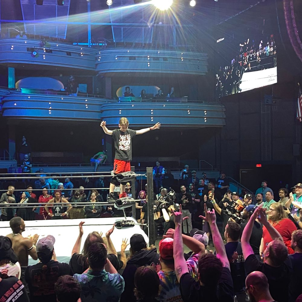 brodie lee jr. standing on the second turnbuckle with his head down and arms out in front of wildly cheering fans. joey janela looms in the background