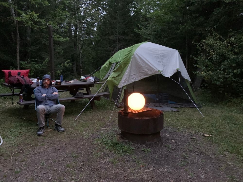 A grumpy looking man sitting in front of a rain fly stretched over a bug screen, next to a fire ring with an LED globe on it