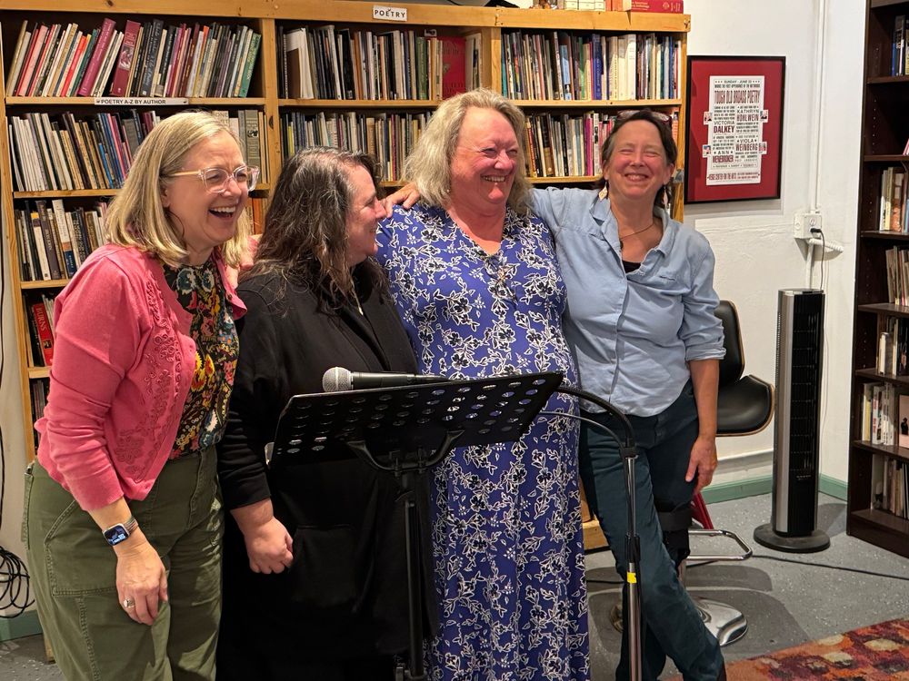 A row of four laughing female poets standing together in front of a wall of books at the Sacramento Poetry Center after their reading. The microphone and black music stand/podium are in front of them.