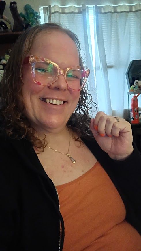 Terra, a trans woman in her late 30s, smiling happily with pink glasses, an orange camisole and gold necklace while wearing a black zip-up jacket.