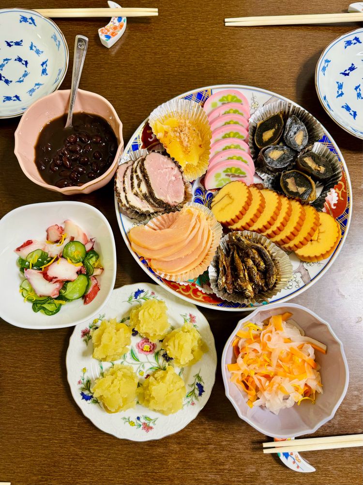 Top down view photo of traditional Japanese new years meal. Counterclockwise: top left bowl of sweet black beans (Kuro-mame), bowl of cucumber & octopus vinegar marinade (su-dako), plate of mashed sweet potatoes with chestnuts (kuri kinton), bowl of julienned daikon radish and carrots vinegar marinade (namasu), and a large platter of peppered ham/kazunoko fish eggs/kamaboko fish cakes/simmered konbu kelp/date-maki egg cake rolls/simmered small fishes/chicken prosciutto