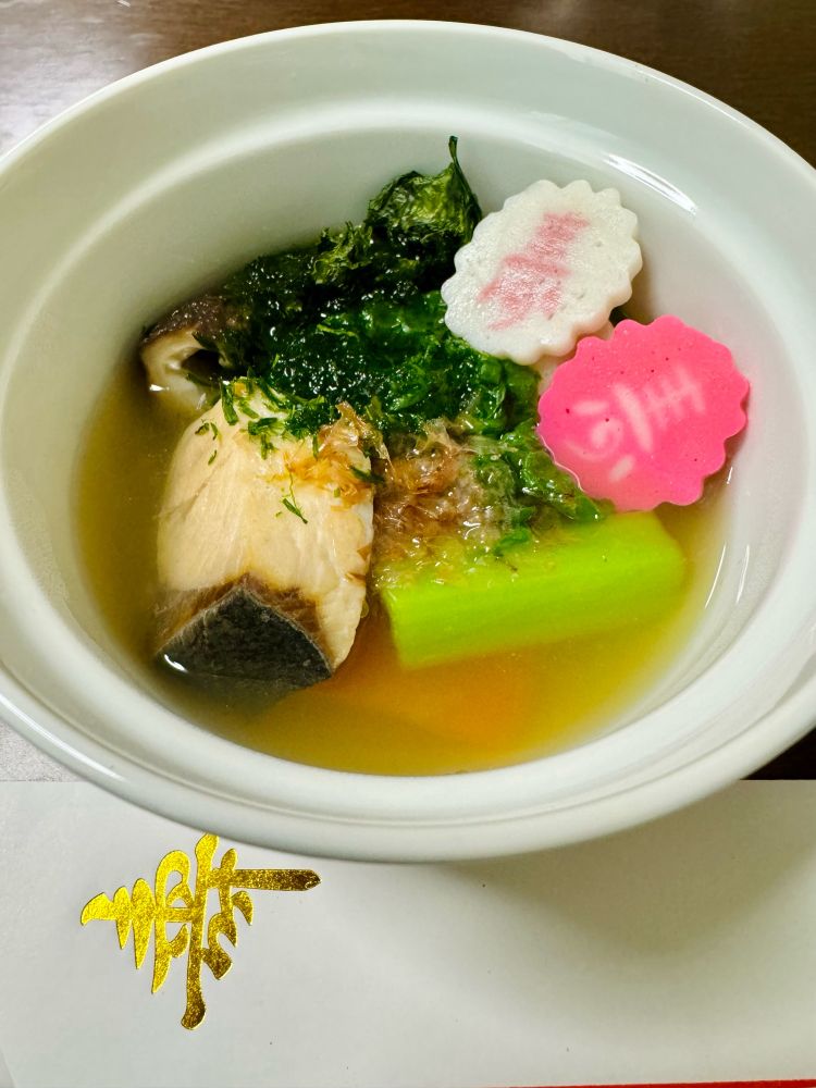 Photo of a bowl of traditional Japanese Zo-ni soup showing: yellowtail, carrots, Katsuo-na greens, red and white fish cake slices, and garnished with dry bonito & aonori seaweed flakes.