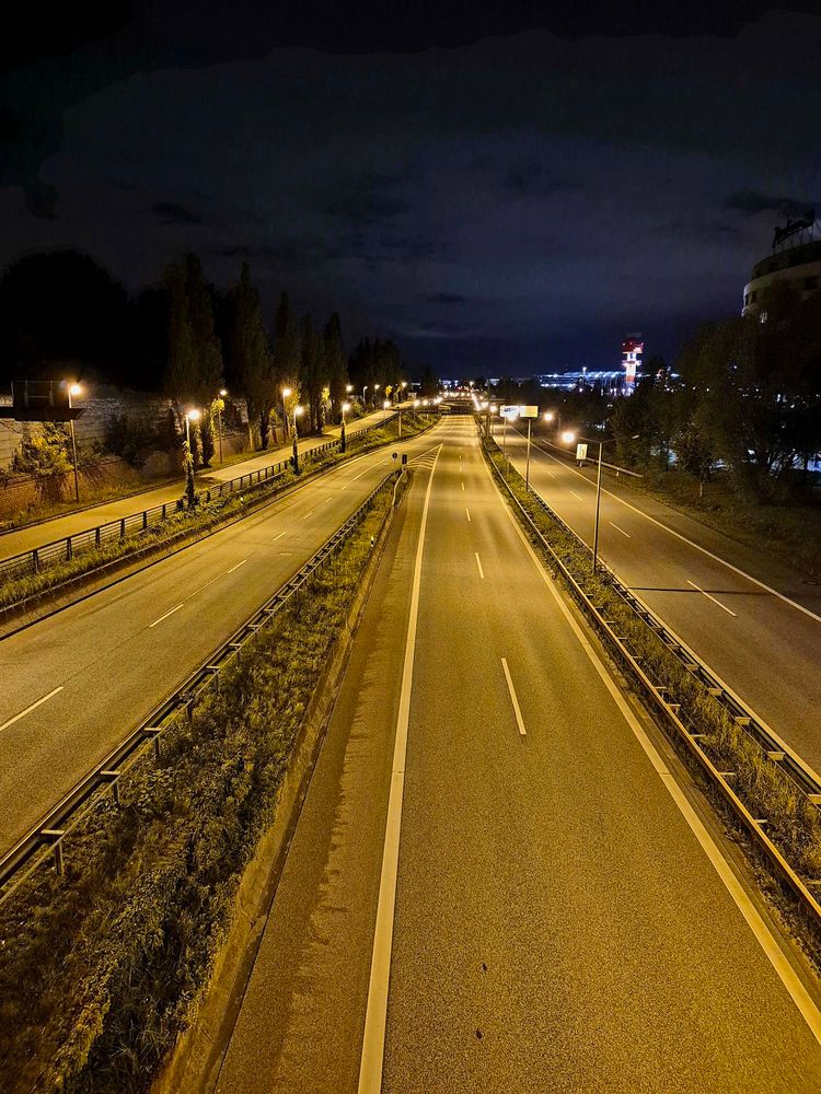 Eine leere Stadtautobahn in der Nacht, die hellerleuchtet ist. In der Ferne sind Gebäude zu erahnen. Der Himmel ist dunkel und zum Teil wolkenverhangen. 