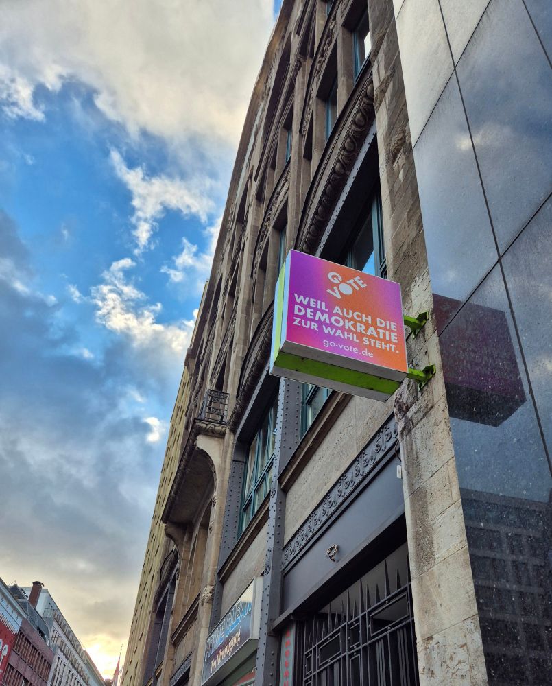 Die Fassade eine Hauses vor bewölktem Himmel. An der Fassade ist ein quadratisches Reklameschild angebracht auf dem steht: go vote, weil auch die Demokratie zur Wahl steht 
