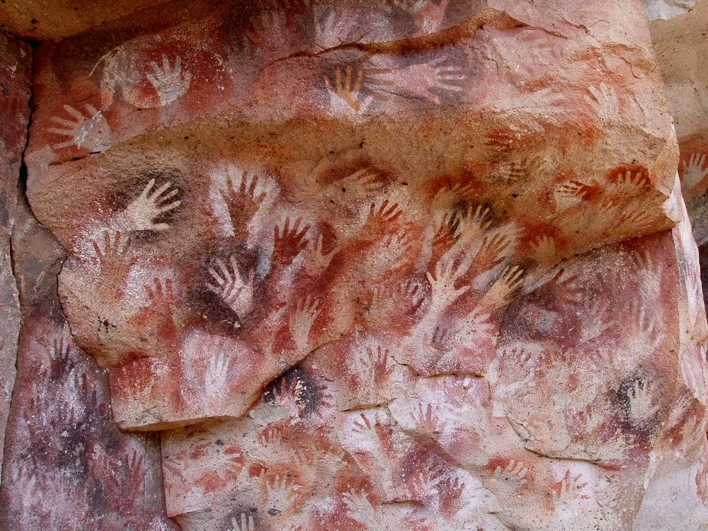 A picture of the handprints at the Cueva de los manos cave in Argentina. The stone wall  is tan and rough. It is a natural stone wall with various ledges and overhangs that eventually transition into the rough ceiling of the cave. On the stone wall are a series  of handprints painted in or outlined in 3 colors: black, white or reddish orange. The hands are numerous, there are so many prints that the newer ones overlap and cover some of the older ones. The handprints have been dated to be from 7500 BC to 700AD. End of transcription.