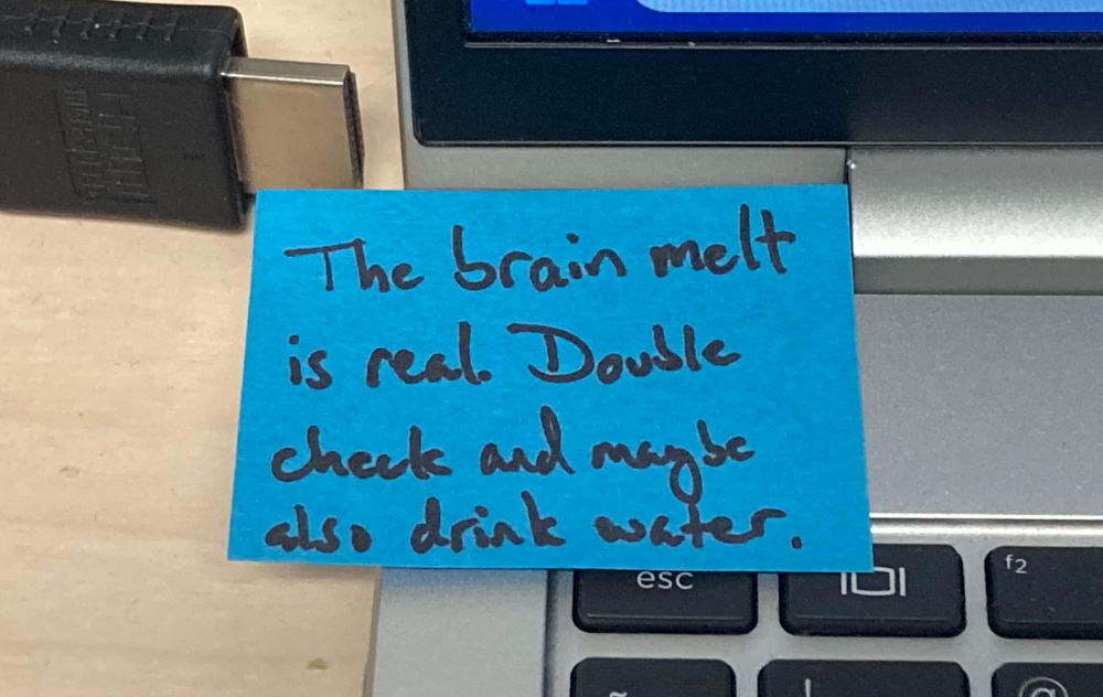 A bright blue mini sticky note on the corner of a laptop between the screen and keyboard. Written on it in black marker is the admonishment “The brain melt is real. Double check and maybe also drink water.” 