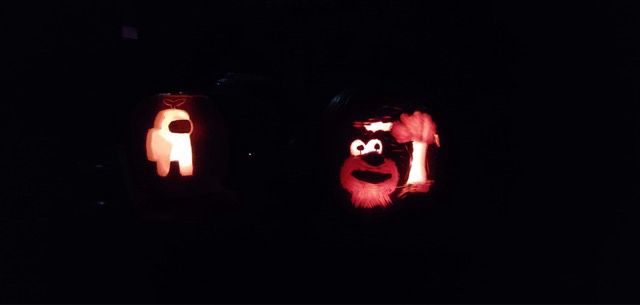 Dark photo of two glowing jack-o-lanterns. On the left is an Among Us crewmate with the plant sprout on its head. On the right is the meme of Elmo looking awed in front of a mushroom cloud. Admittedly, in this carving the mushroom cloud looks more like a broccoli stalk. 
This was my front porch on Halloween in 2020.