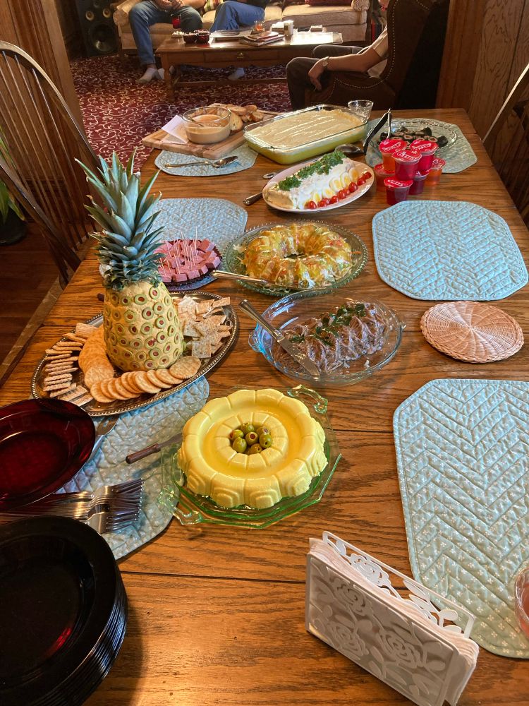 A wooden dining table with quilted place mats, a stack of red glass plates, and an assortment of serving dishes containing mostly gelatinized foods. There’s a bright yellow ring mold with stuffed olives in the center, a fancy log of shredded beef, another ring whose translucence reveals shredded vegetables, a mayonnaise-white log garnished with slices of boiled egg and tomato, some other nondescript casserole dishes at the very end, and a stack of single serving Jello cups. The cheese ball described in the post stands on one edge of the table, decorated with the top of a real pineapple and surrounded by crackers. 